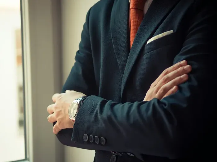 Confident man in a dark suit and orange tie standing by a window with folded arms, symbolizing success on Town Service Spotlight